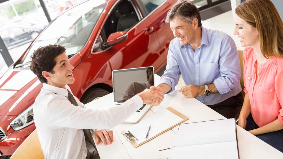 Ein Autoverkäufer schüttelt den Kunden die Hand in einem Autohaus in Cloppenburg, im Hintergrund ist ein rotes Auto zu sehen und auf dem Tisch liegen Unterlagen.